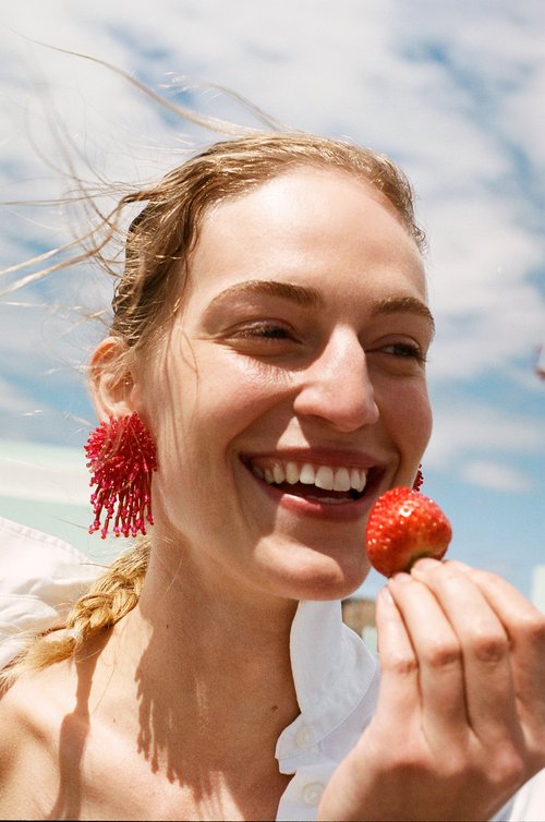 Smiling woman in beaded earrings holds a strawberry against a blue sky with clouds. Fashion and beauty shot with natural lighting.