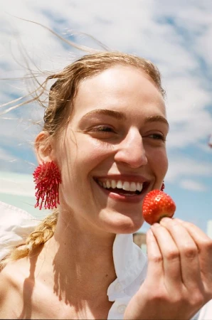 Smiling woman in beaded earrings holds a strawberry against a blue sky with clouds. Fashion and beauty shot with natural lighting.