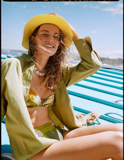 A smiling adult woman in a patterned bikini, green linen shirt, and yellow hat sits on a turquoise surface under a clear sky.
