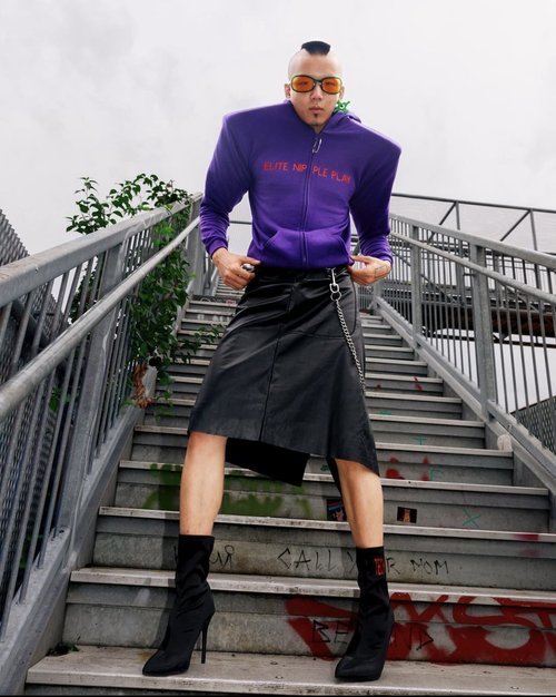 Person on stairs wearing a purple hoodie, black skirt, and black boots poses for a low-angle shot with greenery in the background.