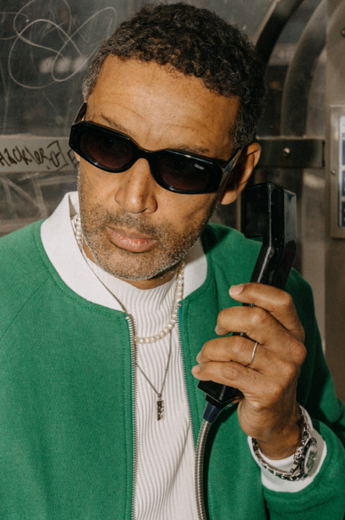 Close-up of an adult man wearing sunglasses and a green bomber jacket, holding a black telephone receiver in an indoor urban setting.