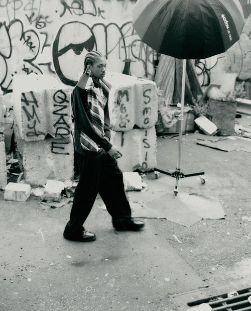 Black and white portrait of a person walking in an urban environment near graffiti-covered blocks, with an umbrella visible overhead.