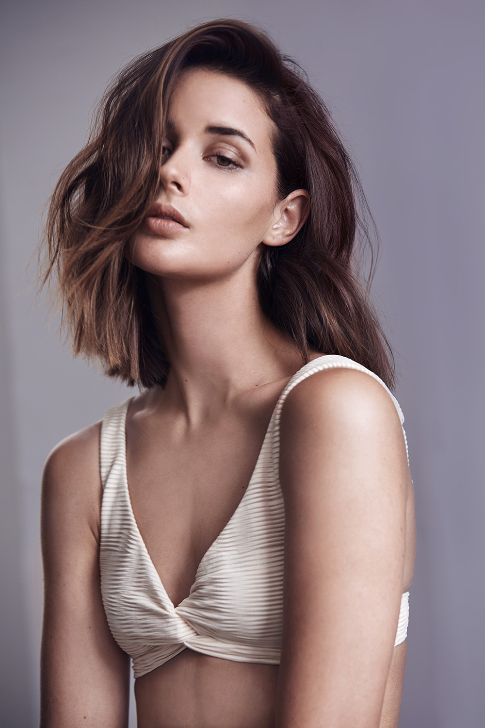 Portrait of a woman with shoulder-length brown hair and a white bikini top against a grey backdrop in a studio setting.