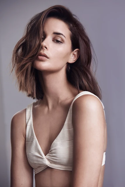 Portrait of a woman with shoulder-length brown hair and a white bikini top against a grey backdrop in a studio setting.