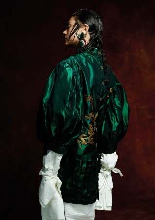 Back view of a woman with braided hair and wearing a green silk blouse, gold earrings, and white trousers against a dark background.