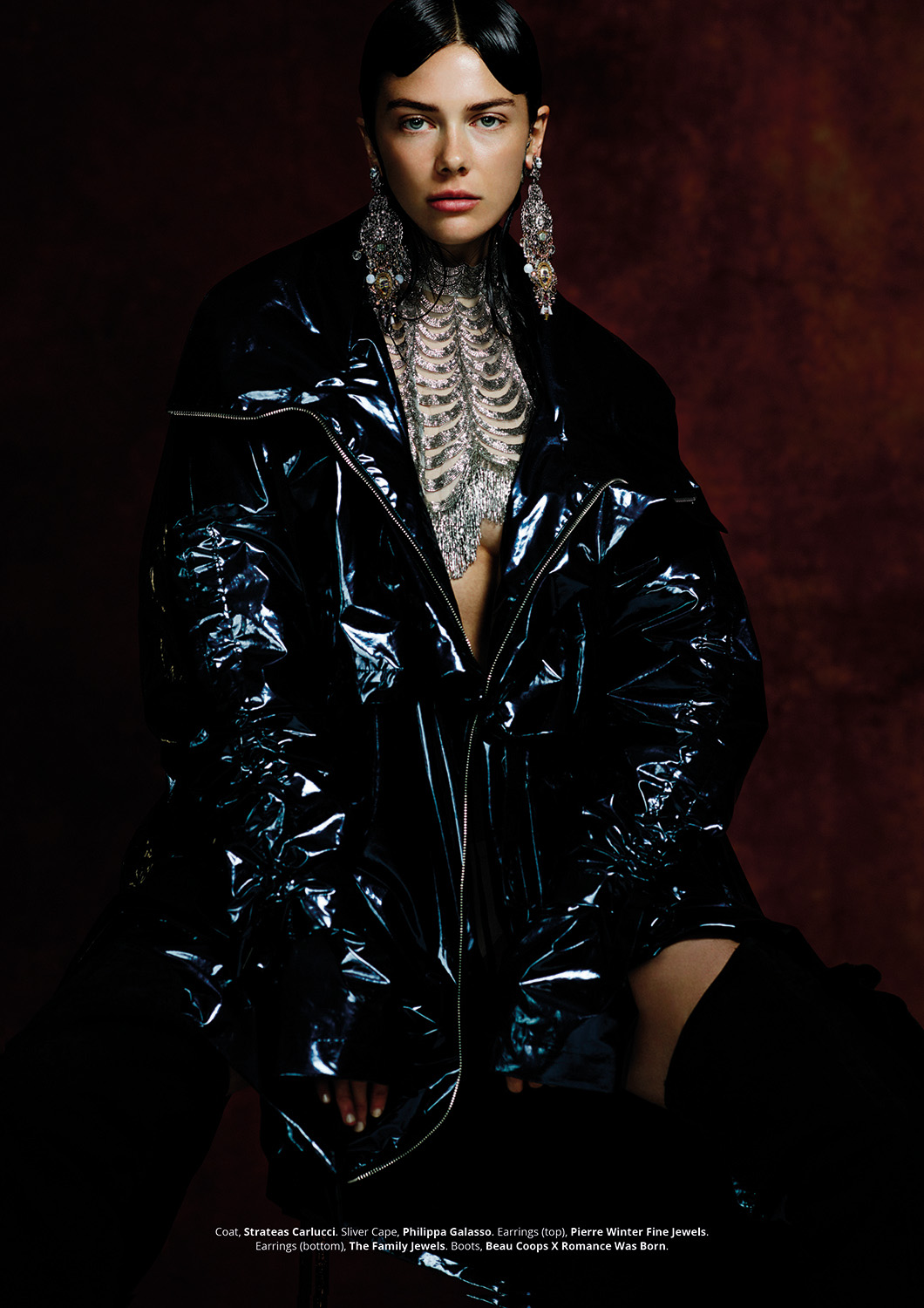 Fashion portrait of a woman wearing a reflective black jacket with an ornate silver necklace and earrings against a brown background.