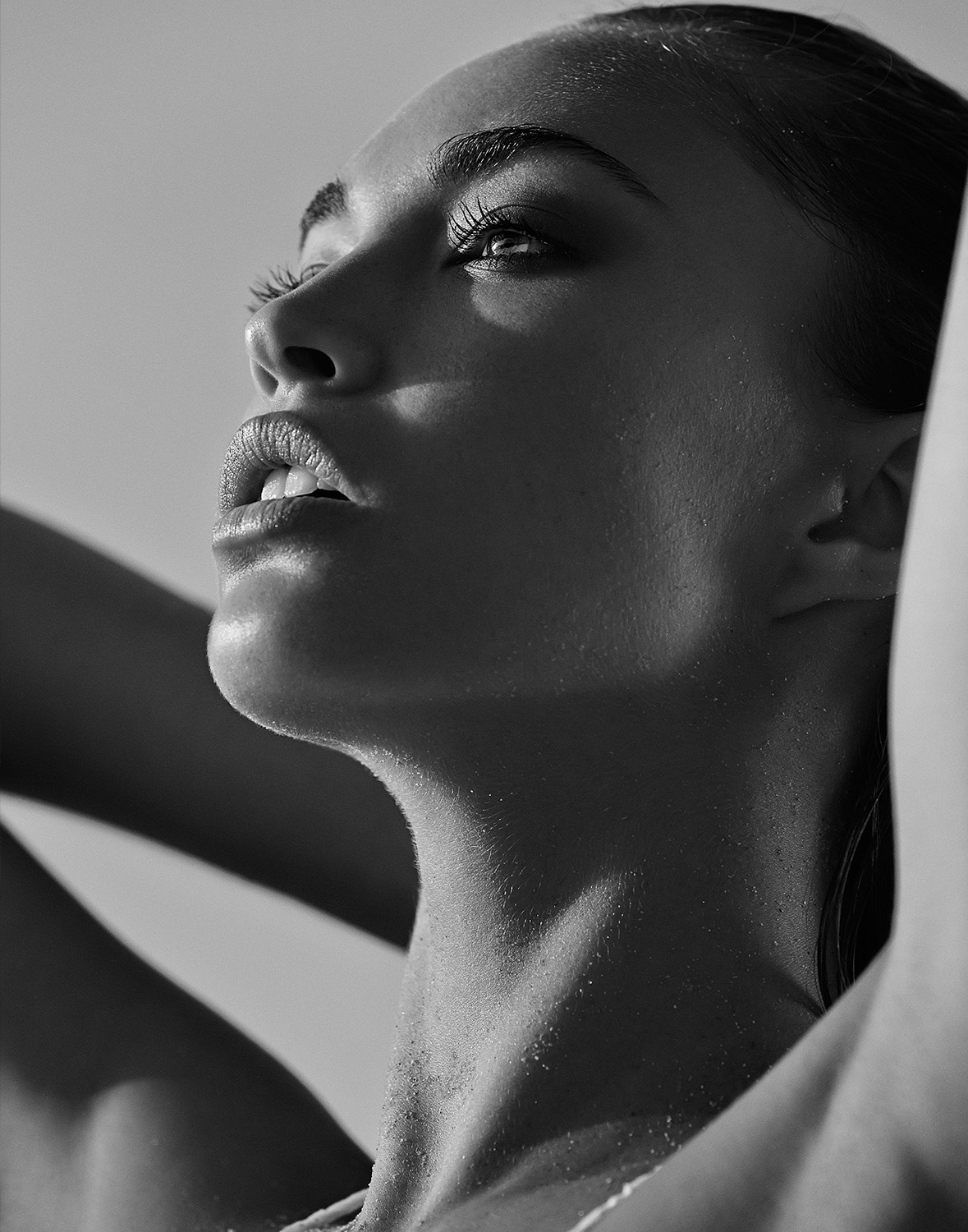 Close-up portrait of a woman looking up with her hands behind her head, black and white photography, bright light source.