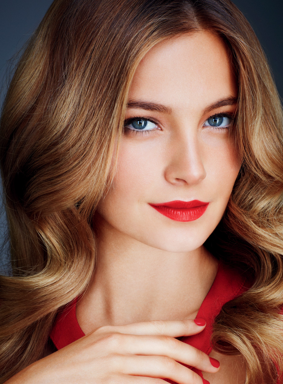 Close-up studio portrait of a woman with wavy blonde hair, red lipstick and a red dress against a dark gray background.