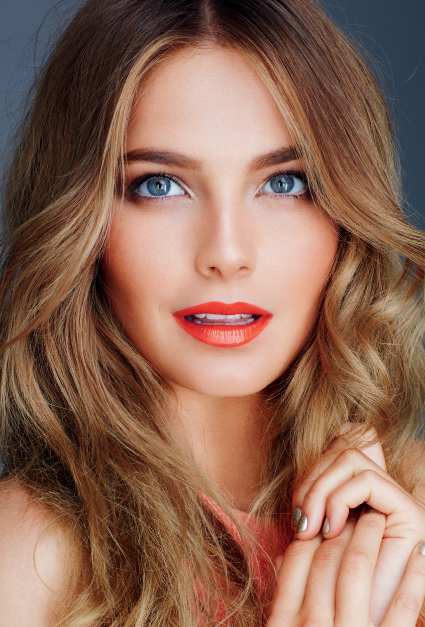 Close-up portrait of a woman with blonde wavy hair, blue eyes, and orange lipstick, posing with her hands near her face.
