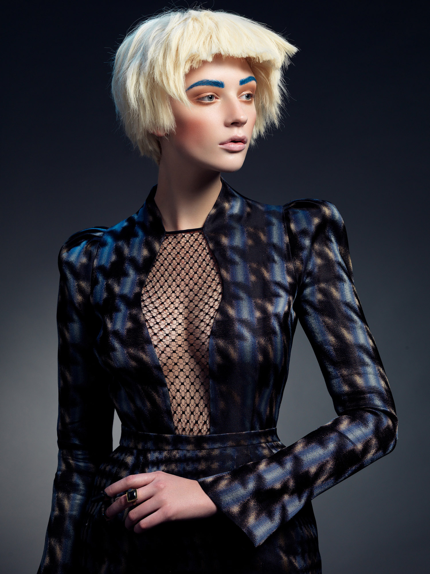 Portrait of a blonde woman with blue eyebrows wearing a blue patterned jacket against a gray background in a beauty editorial fashion shot.