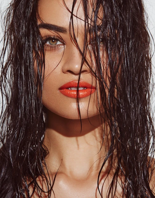 Close-up portrait of a woman with wet dark hair and orange lipstick against a white background in beauty shot with dramatic lighting.