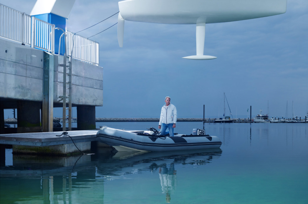 Woman stands in a grey inflatable boat docked by a pier; a large white sculpture sits above. Calm turquoise water and cloudy skies.
