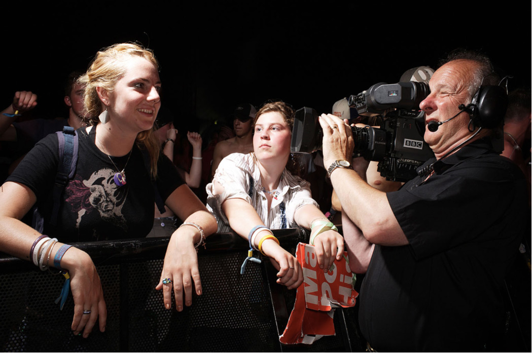 Cameraman films a concert audience with two fair-skinned adult females leaning on a barrier in the foreground at an outdoor event.