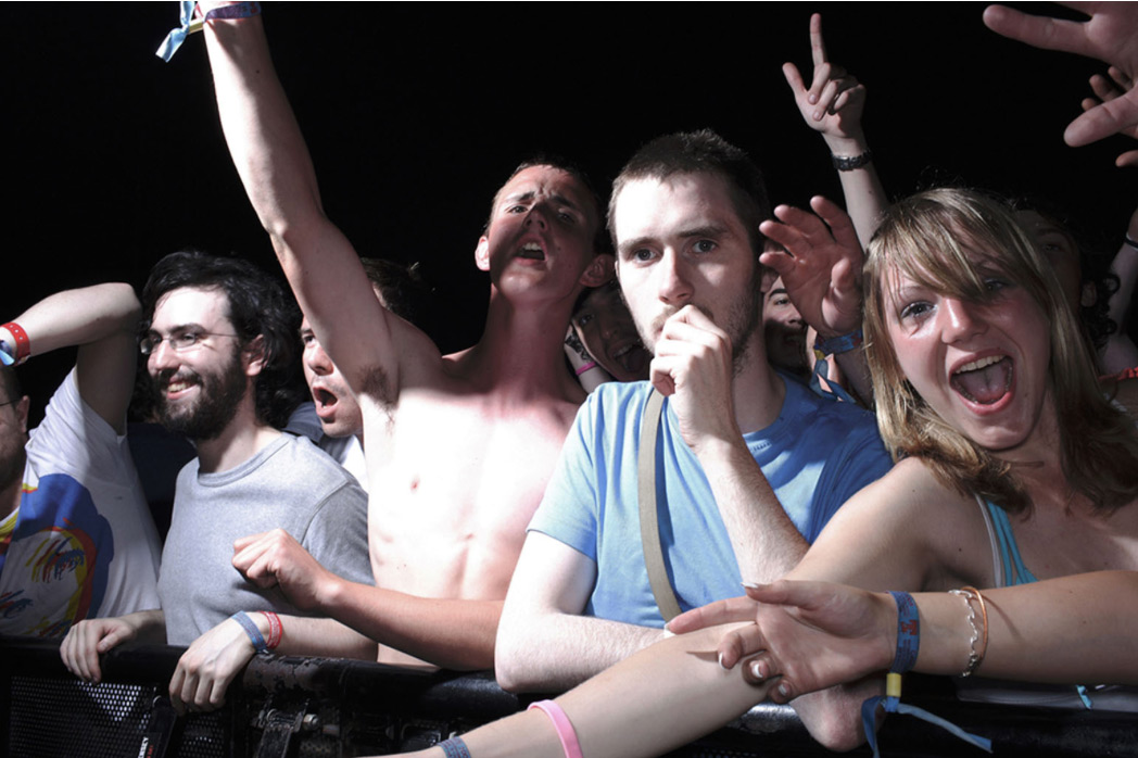 Enthusiastic music fans at an outdoor concert, cheering, with visible wristbands in a dark environment with stage lighting.
