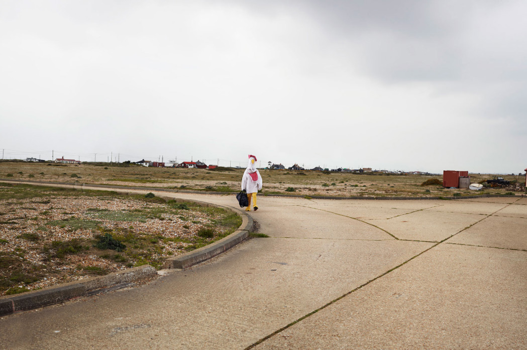Person in a chicken costume walks along an empty road with buildings in the distance under an overcast sky; holding a black bag.