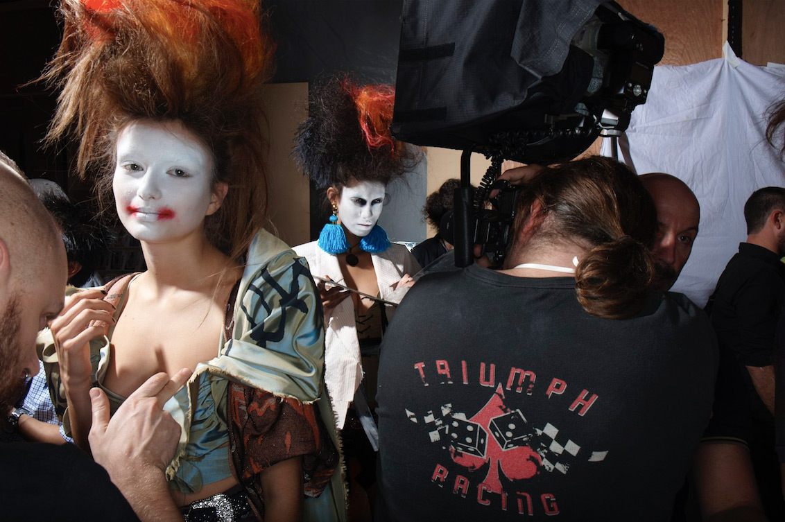 Backstage view of two fashion models with white face makeup and elaborate hair, surrounded by crew and equipment in a photo studio setting.