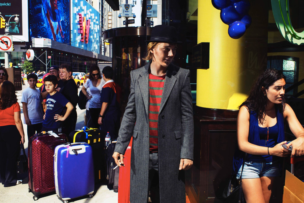 Man in gray coat, black hat stands near luggage in urban scene; other pedestrians, buildings, yellow column visible in the background.