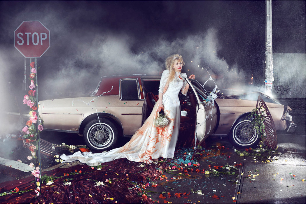 A woman in a wedding dress stands by a damaged vintage car amidst smoke, flowers, and debris in an urban setting, evoking drama and artistry.