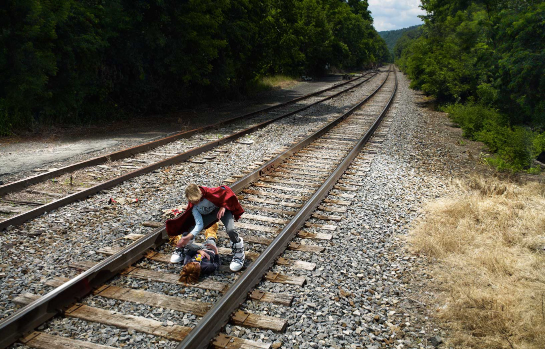 Two children play on train tracks in a rural setting. One child is lying on the tracks while the other kneels beside them in a red cape.