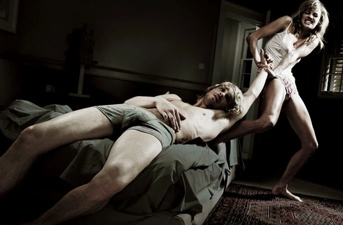A man lounges in bed as a woman aggressively pulls him up by the arm in a dimly lit room with a bed and minimal decor.