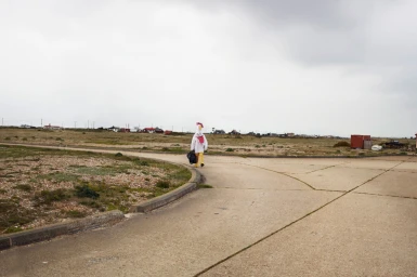 Person in a full-body chicken costume carrying a dark bag walking along a paved road under an overcast sky.