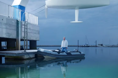 Man standing on an inflatable RIB boat docked near a concrete pier with a large sailboat hull suspended overhead.