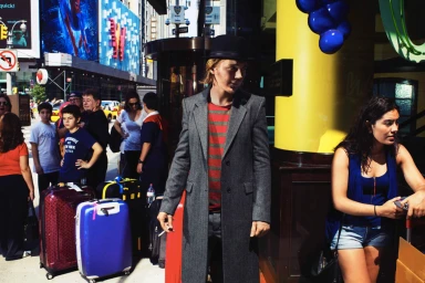 Man in a grey coat and bowler hat standing among luggage on a busy city street with other pedestrians.