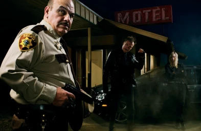 Low-angle shot of a sheriff holding a handgun in the foreground with two figures and a motel exterior at night.