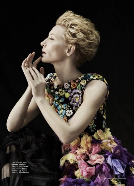 Adult female fashion model with blonde hair poses in profile indoors, wearing a colorful floral dress against a black background.