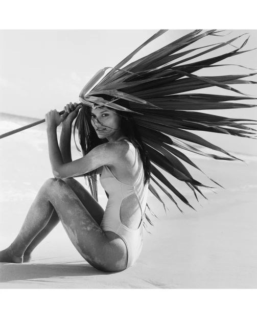 Adult woman in a swimsuit sits on a sandy beach holding large palm fronds in a black and white photograph with a minimalist aesthetic.