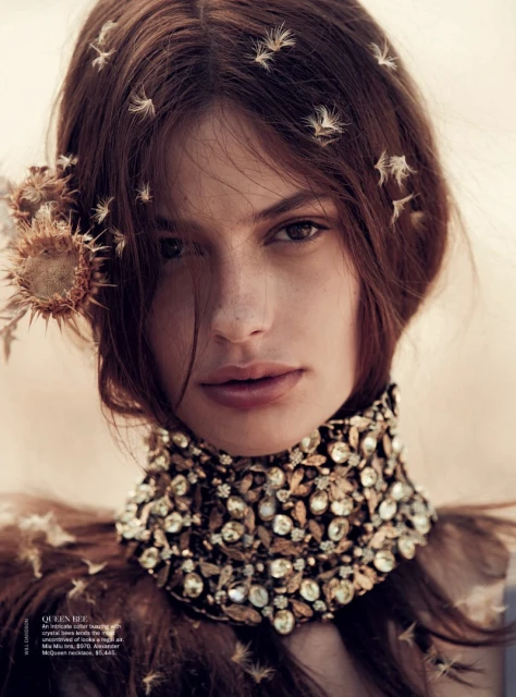 Portrait of a woman with reddish-brown hair wearing an ornate, jeweled collar and dried floral hair accessories in a warm-toned setting.