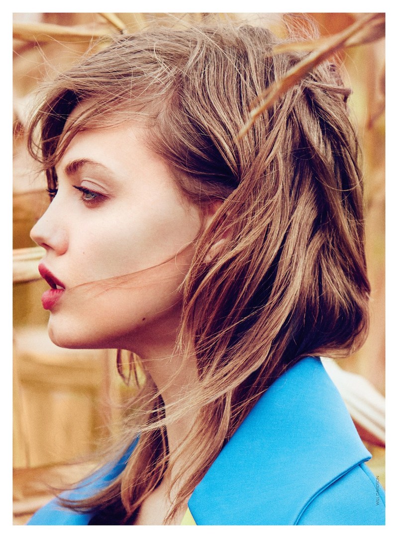Close-up profile of a woman with wavy brunette hair and red lipstick, wearing a turquoise coat, posing in a corn field.