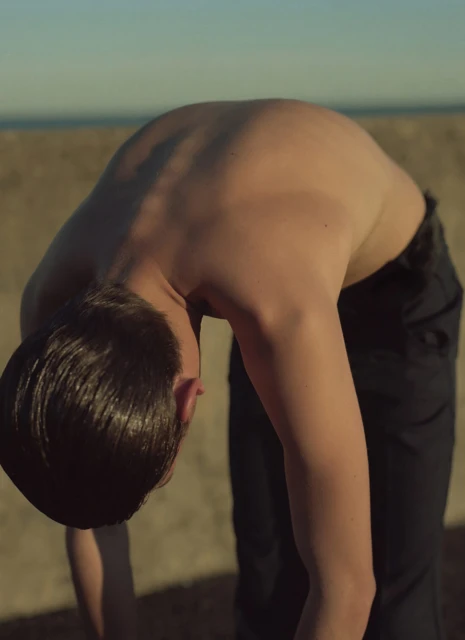A shirtless adult male is bending over with black pants on a sandy beach with a blue sky in the background on a sunny day.