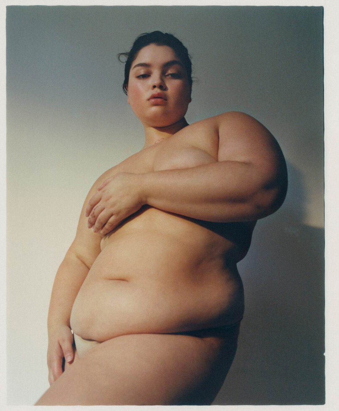 Portrait of a nude woman with dark hair and a fair complexion covering her chest with her right hand, posing against a plain wall.