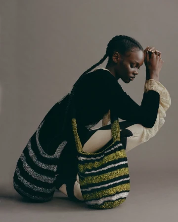 Black woman with braided hair crouching in profile wearing a black top and holding two striped knit shoulder bags