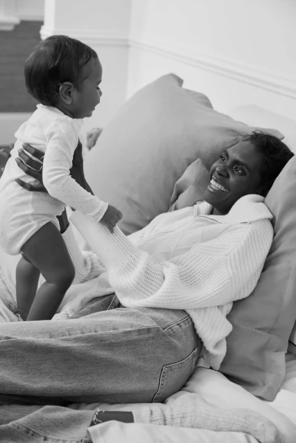 Monochrome image of a mother lying on a bed smiling at her baby, who she is holding up in the air, creating a heartwarming, intimate moment.