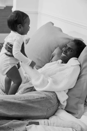 Monochrome image of a mother lying on a bed smiling at her baby, who she is holding up in the air, creating a heartwarming, intimate moment.