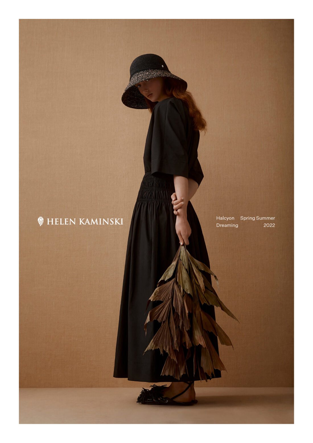 Woman in black dress and hat holding dried leaves, posing in front of warm beige backdrop for Helen Kaminski spring-summer fashion ad.