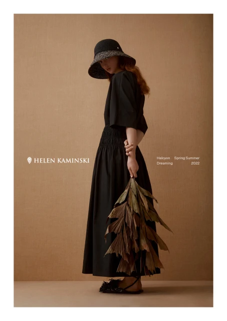 Woman in black dress and hat holding dried leaves, posing in front of warm beige backdrop for Helen Kaminski spring-summer fashion ad.