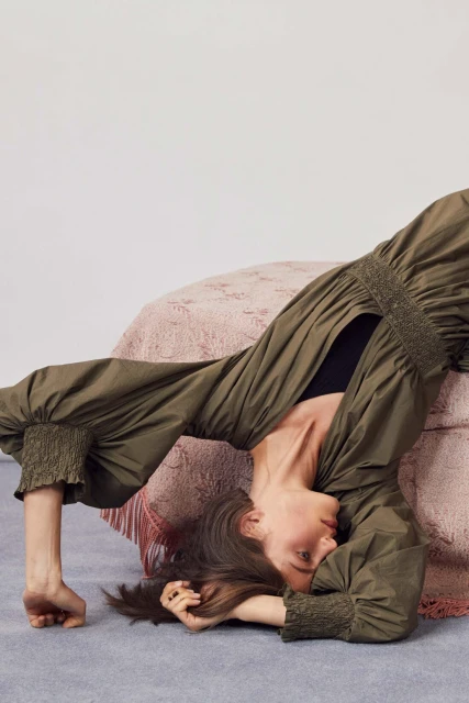 Fashion shot of woman in green dress in yoga pose indoors, with light pink and white color scheme, emphasizing clothing and pose.