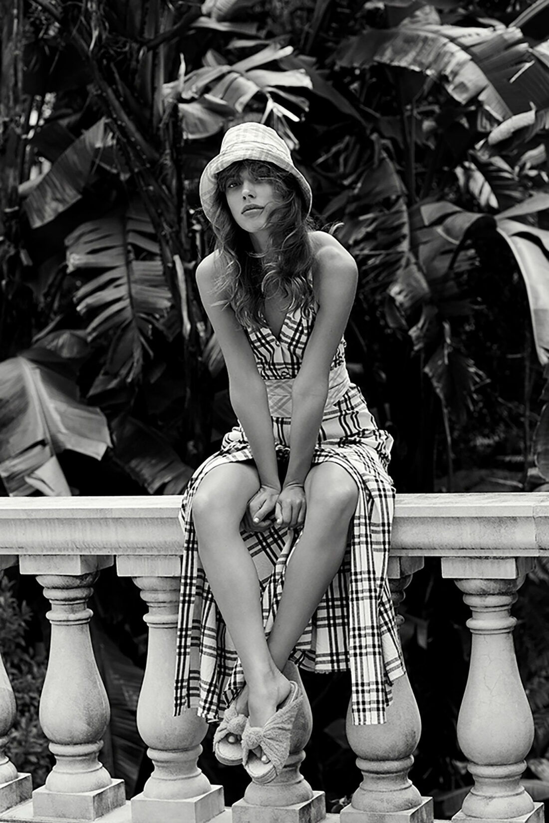 Black and white portrait of a young woman in a plaid dress and hat sitting on a stone railing with lush tropical foliage behind her.