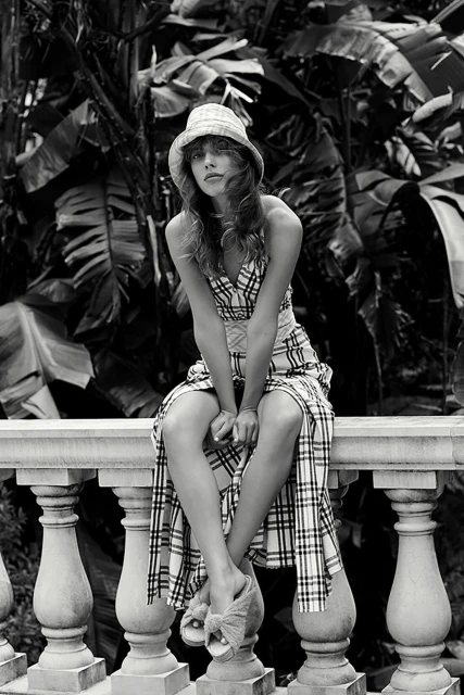 Black and white portrait of a young woman in a plaid dress and hat sitting on a stone railing with lush tropical foliage behind her.
