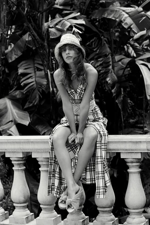 Black and white portrait of a young woman in a plaid dress and hat sitting on a stone railing with lush tropical foliage behind her.