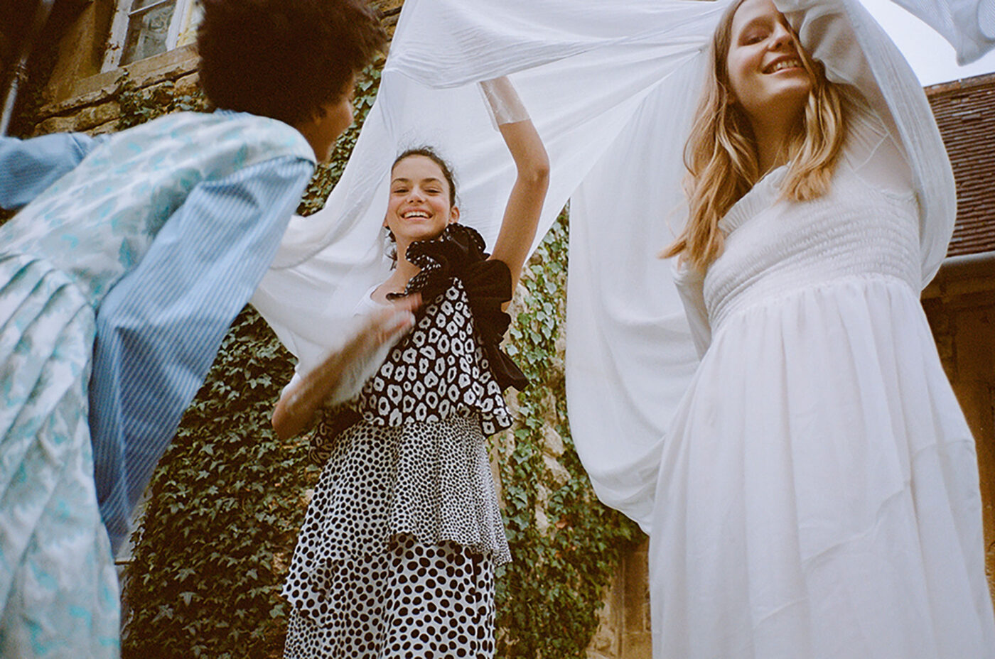 Three young women smiling outdoors, standing near a brick building covered in ivy, wearing dresses and holding up a white sheet.