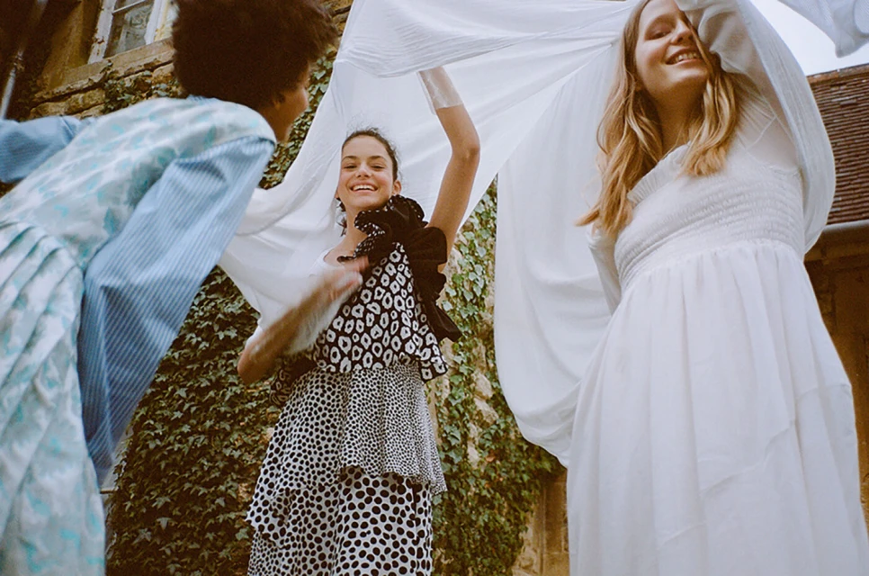 Three young women smiling outdoors, standing near a brick building covered in ivy, wearing dresses and holding up a white sheet.