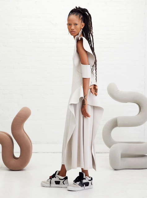 A side profile of a stylish African-American model in an off-white dress with oversized ruffles, white sneakers, and long dreadlocks.