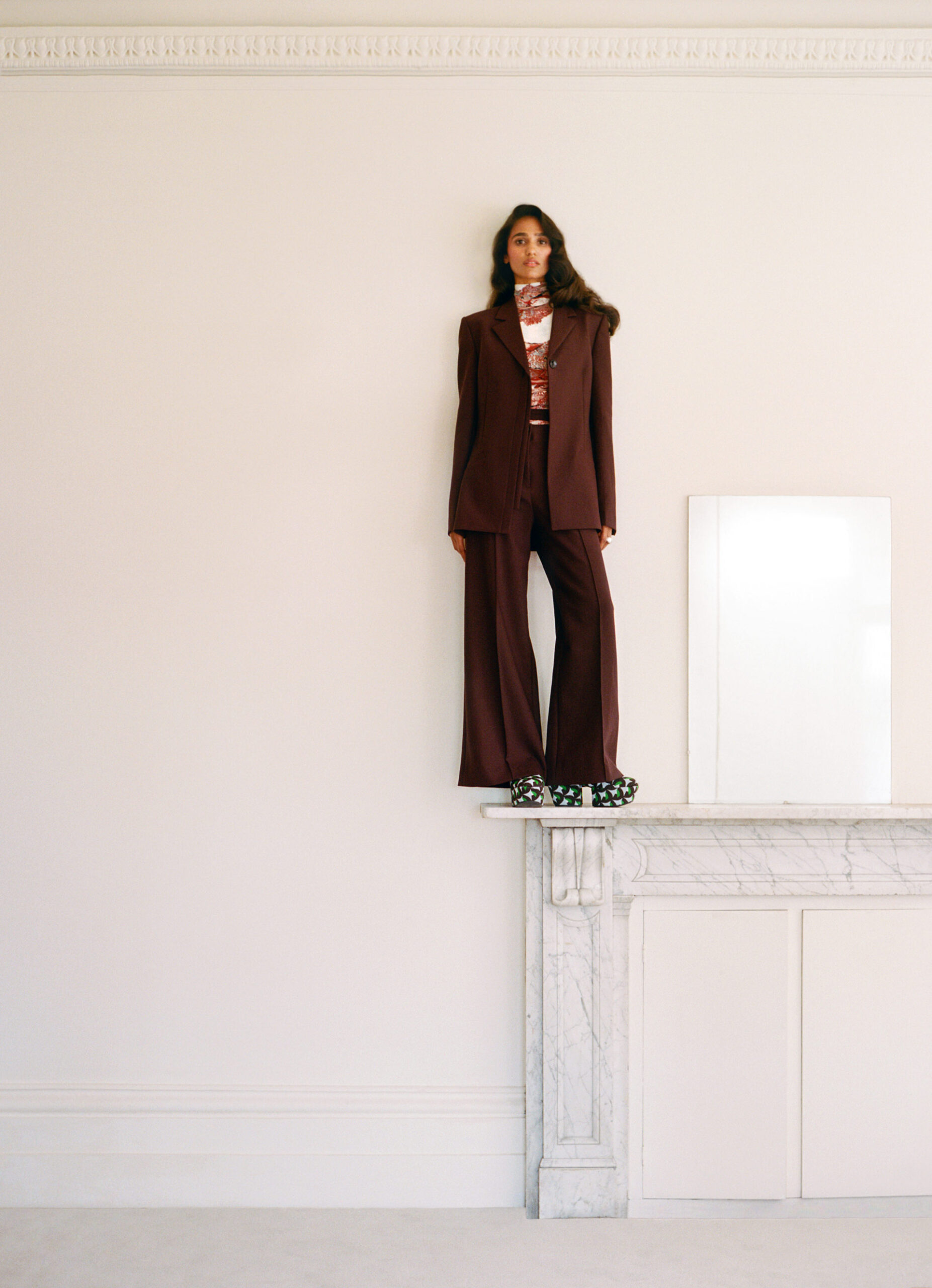 A woman in a brown suit stands on a marble fireplace mantel against a white wall. She is wearing patterned clogs. The setting is indoors.