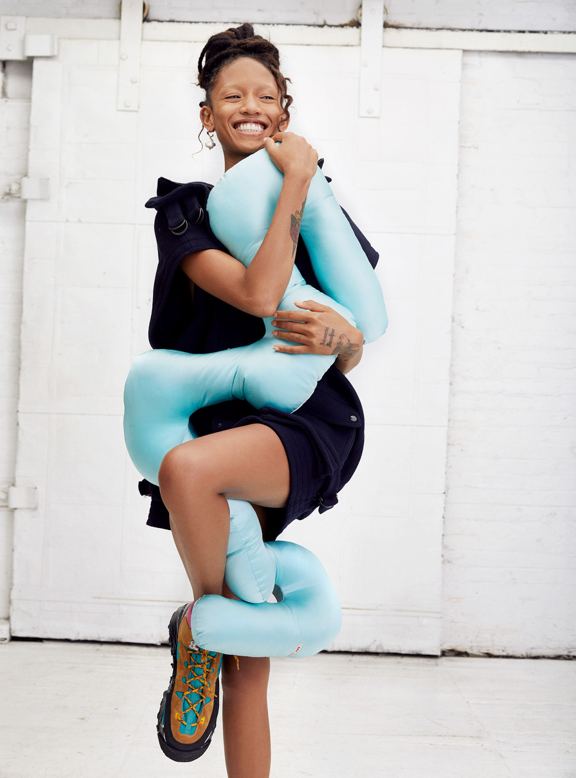 A young adult female model smiles, hugging an aqua-colored plush figure, dressed in a black jacket and dark shorts, set against a white background.