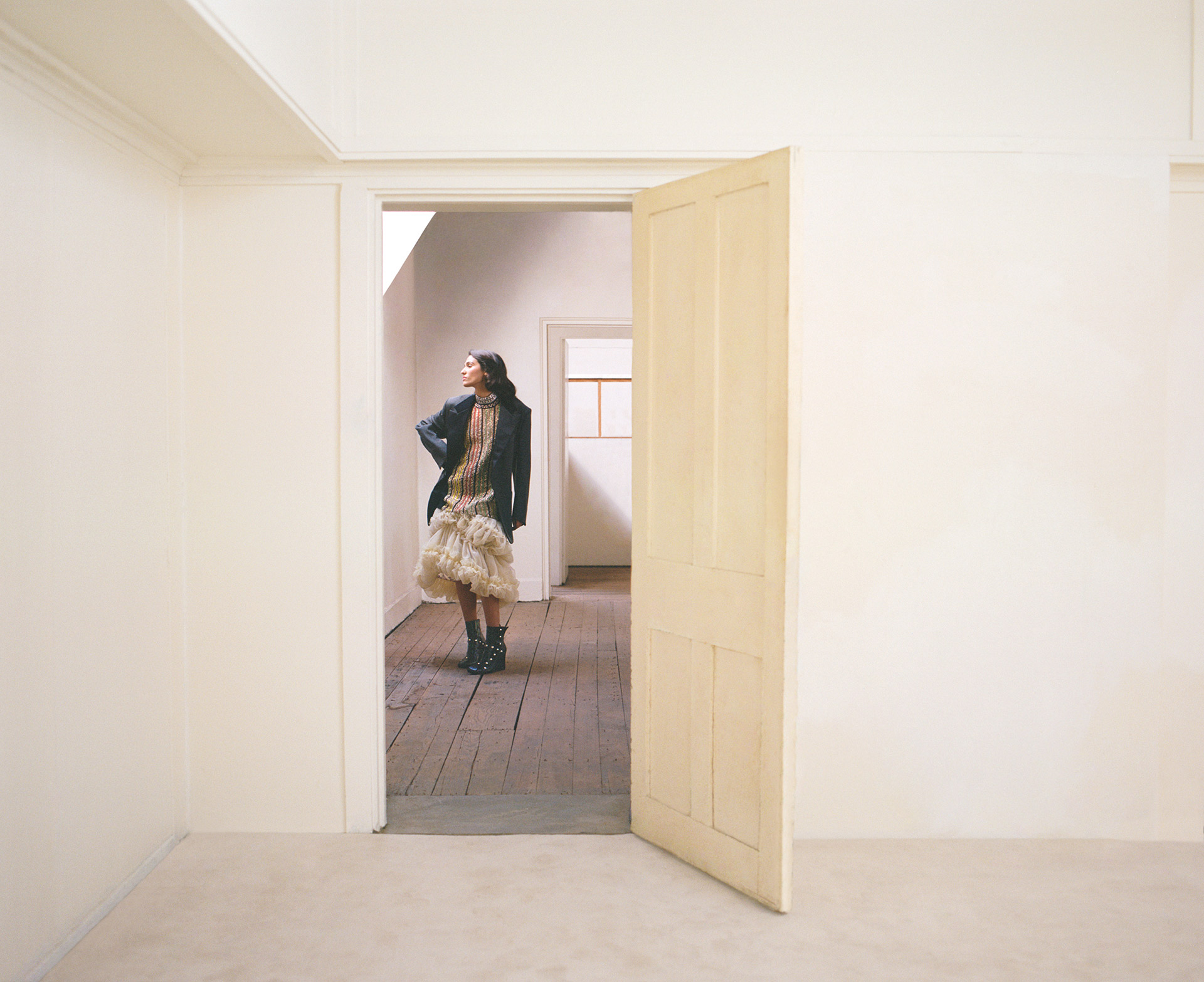 Woman in black jacket and ruffled dress stands in a doorway of a light-filled, minimalistic interior with wood floors.