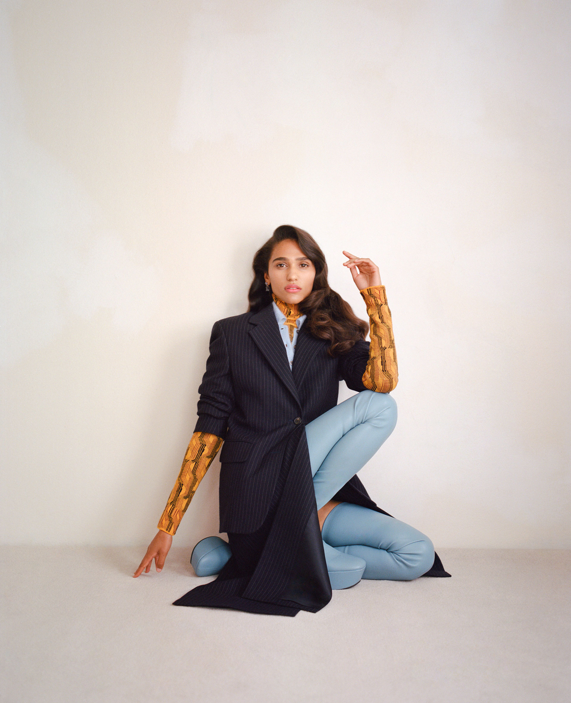 Brunette woman in a pinstripe blazer and light blue pants sits on the floor in a studio, posing with her hand up.